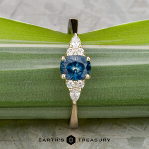 A 14K yellow gold "Arwyn" ring with a 0.95-carat Montana sapphire center, accented by clustered white diamonds, is displayed on a green leaf. "EARTH'S TREASURY" appears at the bottom.