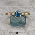 The "Melanie" ring in 18K yellow gold, featuring a 0.94-carat Montana sapphire and diamond-accented sides, is displayed on rough blue crystal above the Earth's Treasury logo.