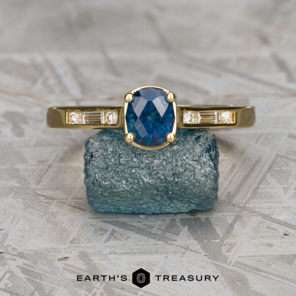 The "Melanie" ring in 18K yellow gold with 0.94-carat Montana sapphire The "Melanie" ring in 18K yellow gold, featuring a 0.94-carat Montana sapphire and diamond-accented sides, is displayed on rough blue crystal above the Earth's Treasury logo.
