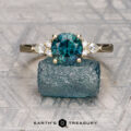 The "Arwyn" ring in 14K yellow gold, featuring a 1.80-carat Montana sapphire and side diamonds, is displayed upright on a rough blue-green stone.