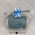 The Cathedral "Lyra" ring in platinum with a 1.44-carat Montana sapphire rests atop a rough blue stone on a textured gray surface, with "Earth's Treasury" labeled at the bottom.