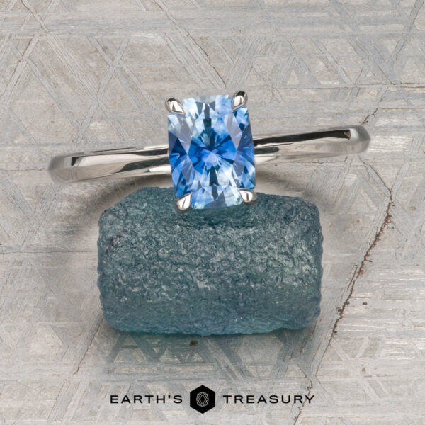 The Cathedral "Lyra" ring in platinum with 1.44-carat Montana sapphire The Cathedral "Lyra" ring in platinum with a 1.44-carat Montana sapphire rests atop a rough blue stone on a textured gray surface, with "Earth's Treasury" labeled at the bottom.