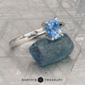 The Cathedral "Lyra" ring in platinum with a 1.44-carat Montana sapphire rests atop a rough blue stone on a textured gray surface, with "Earth's Treasury" labeled at the bottom.