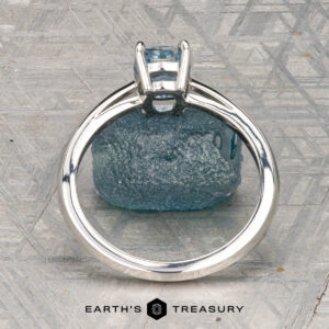 The Cathedral "Lyra" ring in platinum with a 1.44-carat Montana sapphire rests atop a rough blue stone on a textured gray surface, with "Earth's Treasury" labeled at the bottom.