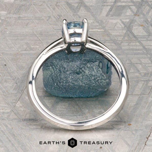 The Cathedral "Lyra" ring in platinum with 1.44-carat Montana sapphire The Cathedral "Lyra" ring in platinum with a 1.44-carat Montana sapphire rests atop a rough blue stone on a textured gray surface, with "Earth's Treasury" labeled at the bottom.