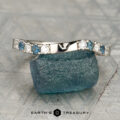 The "Halia" band in 14K white gold, featuring alternating blue and clear gemstones, rests on a rough blue stone atop a textured surface. The Earth’s Treasury logo is displayed at the bottom.