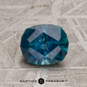 A 1.37-carat deep blue-green Montana Sapphire (heated) is displayed on a textured gray surface with the "EARTH'S TREASURY" logo and text beneath it.