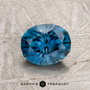 A 1.66-carat peacock blue Montana Sapphire (heated) with intricate faceting sits on a textured gray surface, accompanied by the "EARTH'S TREASURY" text and logo below.