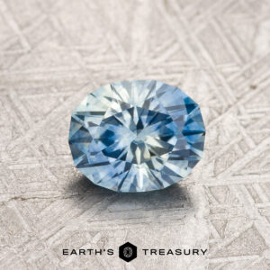 A 1.68-carat Aqua-Medium Blue particolored Montana Sapphire (Heated), brilliant-cut, rests on a textured metallic surface above the "EARTH'S TREASURY" logo.