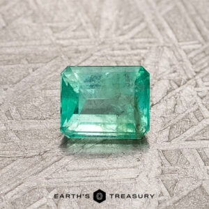 A rectangular, bright green 0.56-carat Colombian emerald with a transparent quality is placed on a textured gray surface, with the text "Earth's Treasury" below.