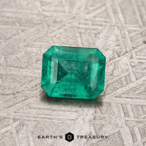 A bright green 0.42-carat Colombian emerald, treated for enhanced beauty, rests on a textured gray surface with the label "Earth's Treasury" below.