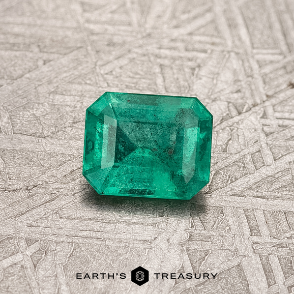 0.42-Carat Colombian Emerald (Treated) A bright green 0.42-carat Colombian emerald, treated for enhanced beauty, rests on a textured gray surface with the label "Earth's Treasury" below.