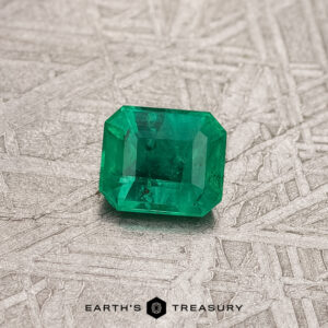 The 0.37-Carat Colombian Emerald (Treated), a vibrant green gem from Colombia, is elegantly showcased on a textured gray surface.