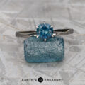 A silver Elenin Solitaire ring, "Nowak, Matthew," with a round blue gemstone stands on a rough blue crystal atop a light textured surface. The "EARTH'S TREASURY" logo is shown at the bottom.