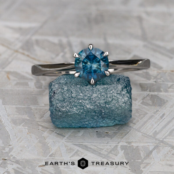 Nowak, Matthew A silver Elenin Solitaire ring, "Nowak, Matthew," with a round blue gemstone stands on a rough blue crystal atop a light textured surface. The "EARTH'S TREASURY" logo is shown at the bottom.
