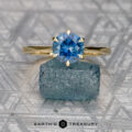 The Raines, Dylan gold solitaire ring with a round blue Elenin gemstone rests on rough blue mineral, set against a geometric patterned background with the "Earth's Treasury" logo below.