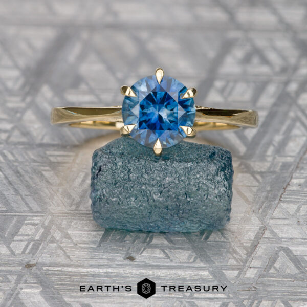 Raines, Dylan The Raines, Dylan gold solitaire ring with a round blue Elenin gemstone rests on rough blue mineral, set against a geometric patterned background with the "Earth's Treasury" logo below.