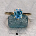 The "Summers, Benjamin (Ben) 1" ring with an oval blue gemstone stands on a rugged blue mineral, set against a geometric backdrop with "EARTH'S TREASURY" below, capturing the elegance of the Elenin collection.