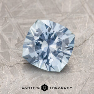 A 2.01-Carat Pale Blue Montana Sapphire rests on a textured, metallic surface. The "Earth's Treasury" logo and text are visible at the bottom.