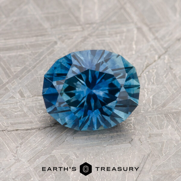 1.42-Carat Peacock Blue Montana Sapphire (Heated) A round, faceted 1.42-Carat Peacock Blue Montana Sapphire (Heated) rests on a textured surface with "EARTH'S TREASURY" and a small geometric logo below.