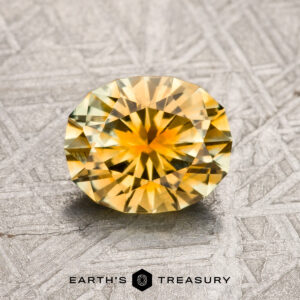 A 1.47-carat gold-green particolored Montana Sapphire (heated) with a round, brilliant cut rests on a textured gray surface; the "Earth's Treasury" logo appears below.
