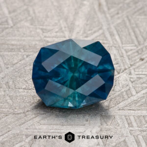 A 1.74-carat Hazy Deep Blue-Green Montana Sapphire (Heated) rests on a textured metallic surface with the "Earth's Treasury" logo and text below.