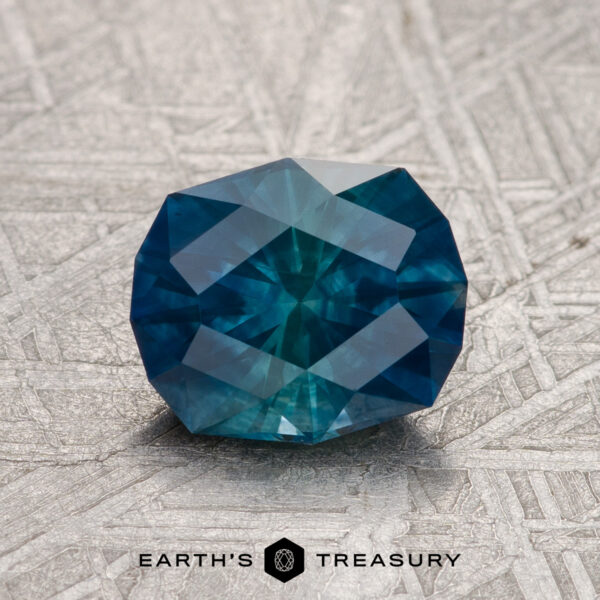 1.74-Carat Hazy Deep Blue-Green Montana Sapphire (Heated) A 1.74-carat Hazy Deep Blue-Green Montana Sapphire (Heated) rests on a textured metallic surface with the "Earth's Treasury" logo and text below.
