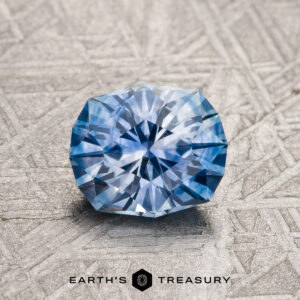 A 1.37-carat sky-pale blue particolored Montana Sapphire (heated), cut in a round brilliant style, rests on a textured gray surface with the "Earth's Treasury" logo and text below.