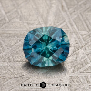 A 1.26-carat blue-green bicolored Montana sapphire (heated), cushion-cut, is shown on a metallic patterned surface with the text "EARTH'S TREASURY" below.