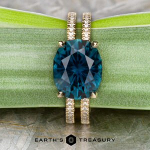 Stacked from 39 images. Method=B (R=8, S=4) gold Gaia ring with large oval blue gemstone and two textured gold bands, displayed on a green leaf. 'Earth's Treasury' logo and text below.