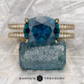 The "Gaia" ring in 14k yellow gold with a 4.63-carat Montana Sapphire and double diamond bands is shown on a rough blue crystal, its elegance highlighted by a gray textured background. "EARTH'S TREASURY" appears at the bottom.
