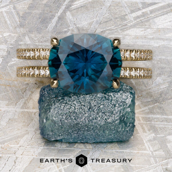 The "Gaia" ring in 14k yellow gold with 4.63-Carat Montana Sapphire The "Gaia" ring in 14k yellow gold with a 4.63-carat Montana Sapphire and double diamond bands is shown on a rough blue crystal, its elegance highlighted by a gray textured background. "EARTH'S TREASURY" appears at the bottom.