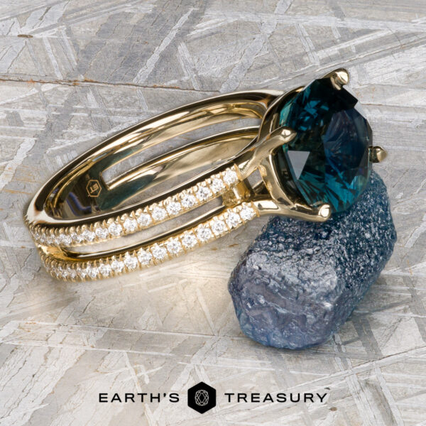The "Gaia" ring in 14k yellow gold with 4.63-Carat Montana Sapphire The "Gaia" ring in 14k yellow gold, featuring a 4.63-carat Montana sapphire and diamond-studded band, rests on a rough stone with the Earth's Treasury logo below.