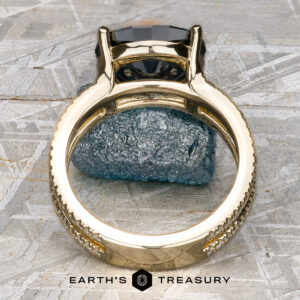 The "Gaia" ring in 14k yellow gold, featuring a 4.63-carat Montana Sapphire, stands upright on rough gray stone, with the Earth's Treasury logo at the bottom of the image.
