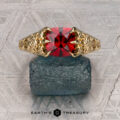 The "Guanyin" ring in 18K yellow gold, featuring a 1.94-carat Malaia garnet, is displayed on dark stone with a geometric patterned background.