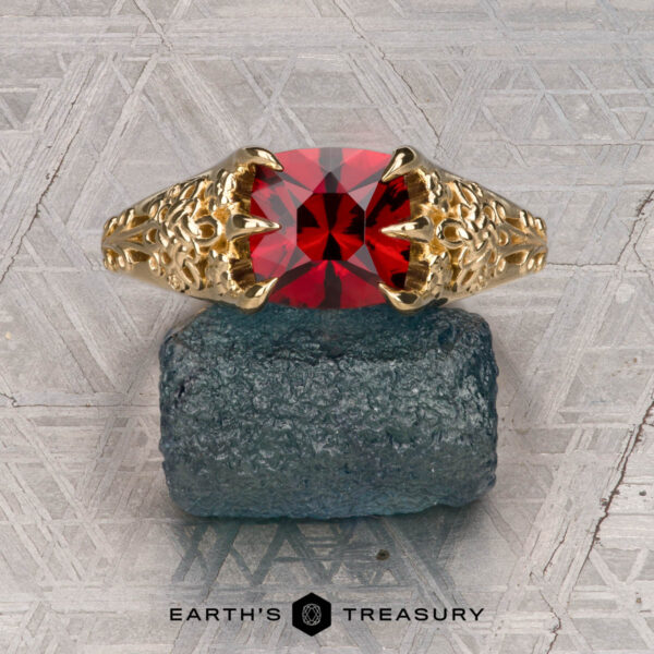 The "Guanyin" ring in 18K yellow gold with 1.94-carat Malaia garnet The "Guanyin" ring in 18K yellow gold, featuring a 1.94-carat Malaia garnet, is displayed on dark stone with a geometric patterned background.