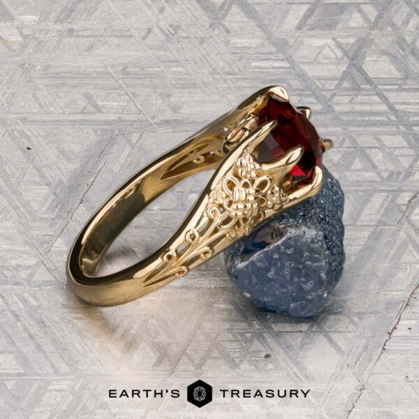 The "Guanyin" ring in 18K yellow gold with 1.94-carat Malaia garnet The "Guanyin" ring in 18K yellow gold, featuring a 1.94-carat Malaia garnet, is displayed on dark stone with a geometric patterned background.