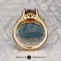 The "Guanyin" ring in 18K yellow gold, featuring a 1.94-carat Malaia garnet, is displayed on dark stone with a geometric patterned background.
