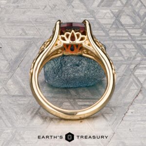 The "Guanyin" ring in 18K yellow gold, featuring a 1.94-carat Malaia garnet, is displayed on dark stone with a geometric patterned background.