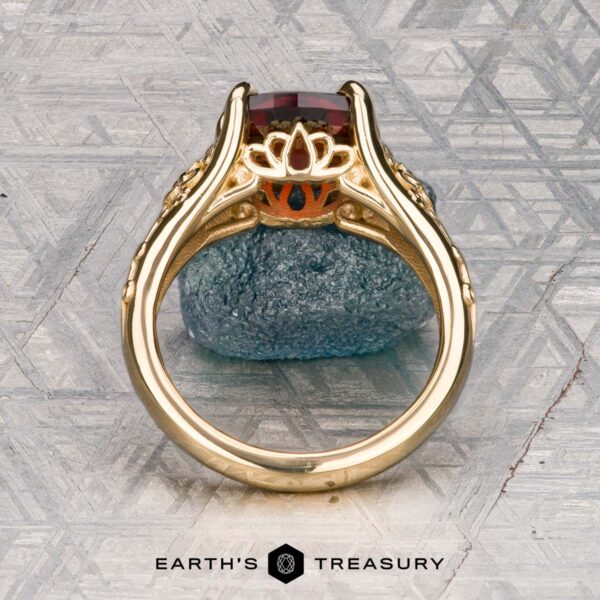 The "Guanyin" ring in 18K yellow gold with 1.94-carat Malaia garnet The "Guanyin" ring in 18K yellow gold, featuring a 1.94-carat Malaia garnet, is displayed on dark stone with a geometric patterned background.