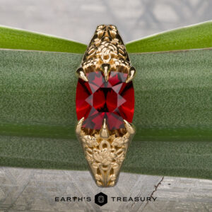 The "Guanyin" ring in 18K yellow gold, featuring a 1.94-carat Malaia garnet, is displayed on dark stone with a geometric patterned background.