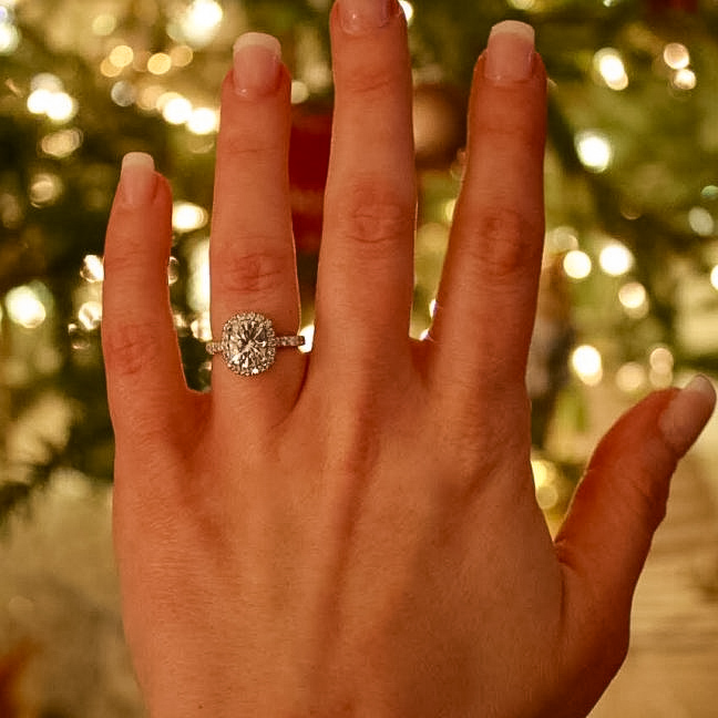 A hand wearing the AC Testimonial-1200 ring, adorned with a large diamond, is pictured against a blurred, illuminated Christmas tree.