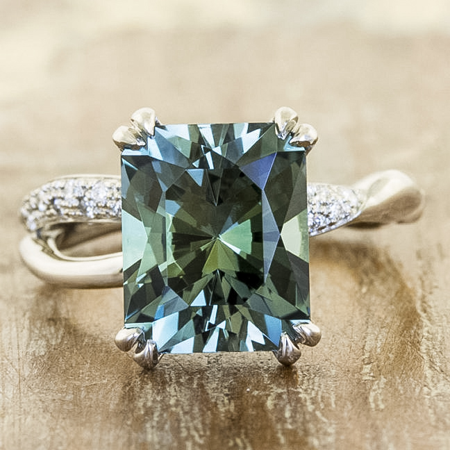 The CG Testimonial-1200 is an emerald-cut green gemstone ring featuring a twisted band with diamond accents, displayed on a wooden surface.