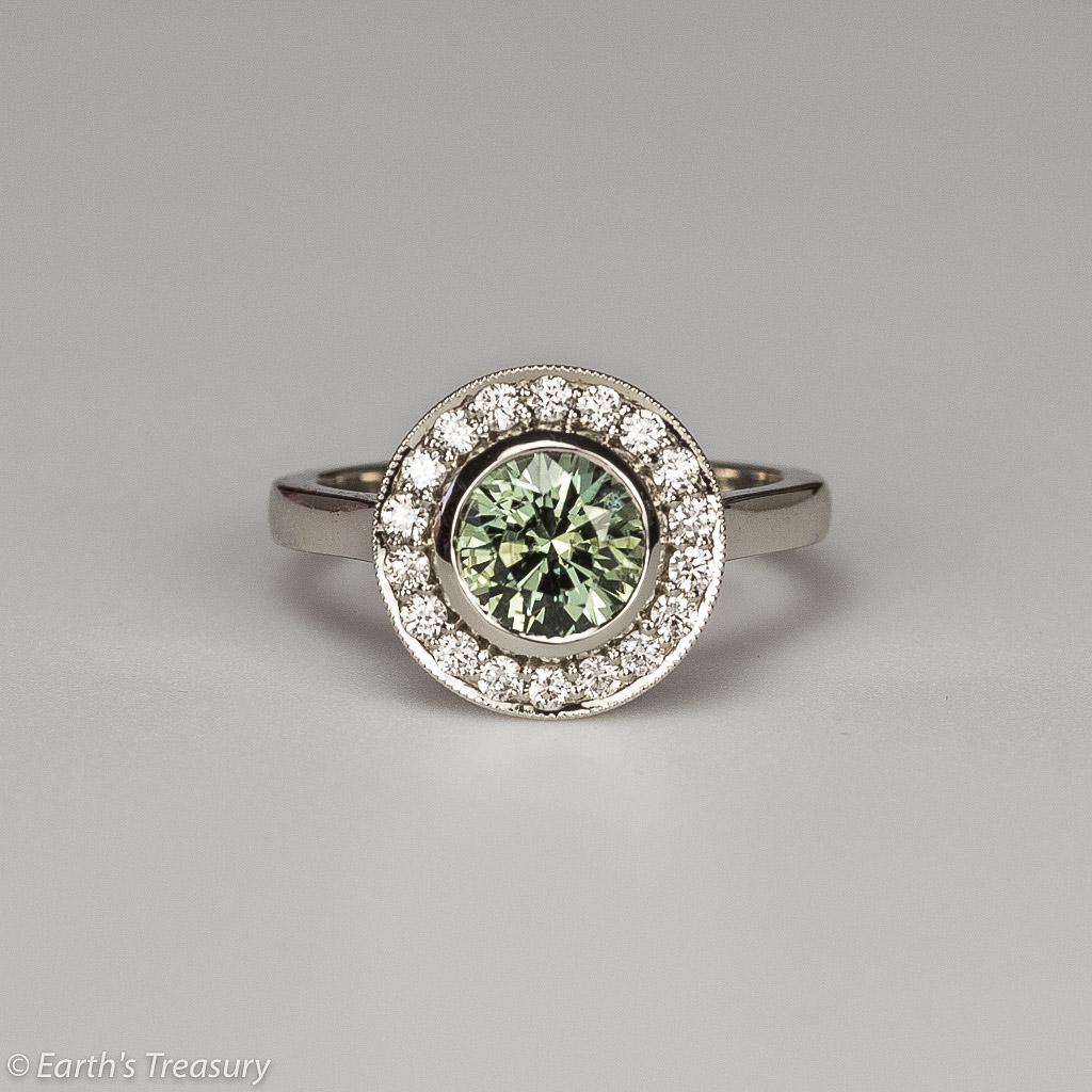 The GN Testimonial-1200 is a silver ring with a central green gemstone encircled by small clear stones in a circular setting, set against a plain background.