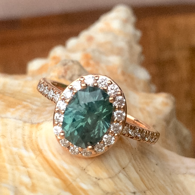 The J Testimonial-1200 is a gold ring featuring an oval green gemstone encircled by small diamonds, showcased on a seashell.