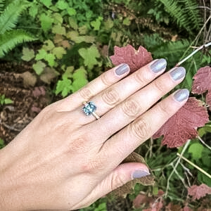 A hand with gray-painted nails wearing the KJ Testimonial-1200, featuring a blue gemstone, is pictured over red leaves in a natural outdoor setting.