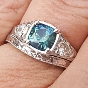 A close-up of a hand wearing the Kelly Testimonial-1200, a silver ring featuring a central square blue-green gemstone with engraved decorative details.