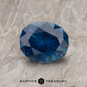 A 1.52-Carat Deep Blue Montana Sapphire (Heated) is shown on a textured gray surface with the "Earth's Treasury" logo at the bottom.
