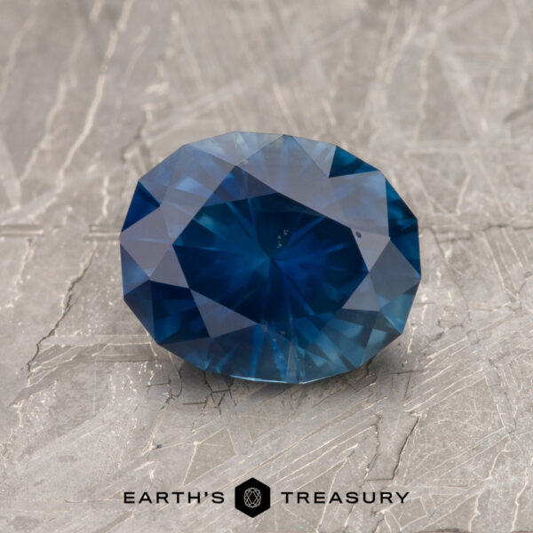 1.52-Carat Deep Blue Montana Sapphire (Heated) A 1.52-Carat Deep Blue Montana Sapphire (Heated) is shown on a textured gray surface with the "Earth's Treasury" logo at the bottom.