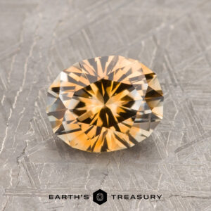 A 1.43-carat orange-colorless particolored Montana Sapphire (heated) rests on a textured metallic surface, with the "Earth's Treasury" logo and text below.